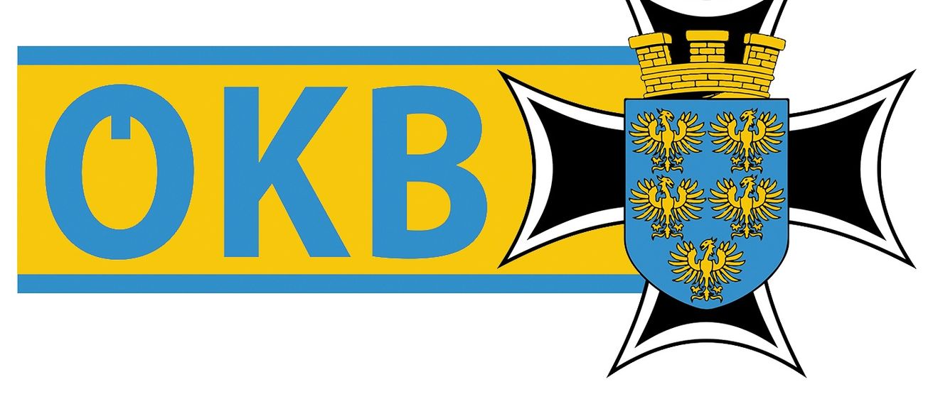 Das Bild zeigt ein heraldisches Emblem mit den Buchstaben 'KB' in großen blauen Buchstaben auf gelbem Hintergrund. Dahinter befindet sich ein schwarzer Kreuz. Das Emblem umfasst einen blauen Schild mit einem goldenen Adler, einer Burg und anderen Symbolen.