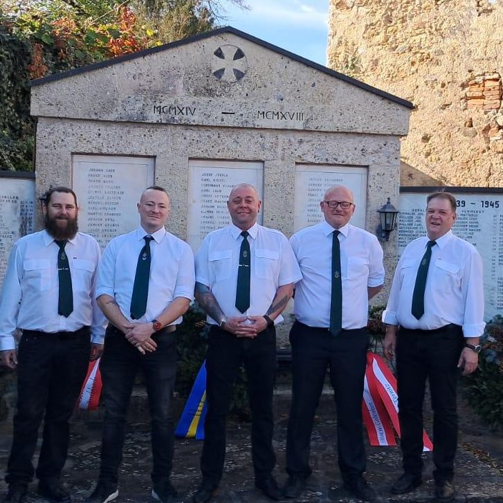 Fünf Männer stehen vor einem Denkmal, gekleidet in weiße Hemden und grüne Krawatten. Hinter ihnen hat eine Steinmauer Tafeln mit Namen und Jahren, die möglicherweise diejenigen ehren, die im Zweiten Weltkrieg gedient haben.