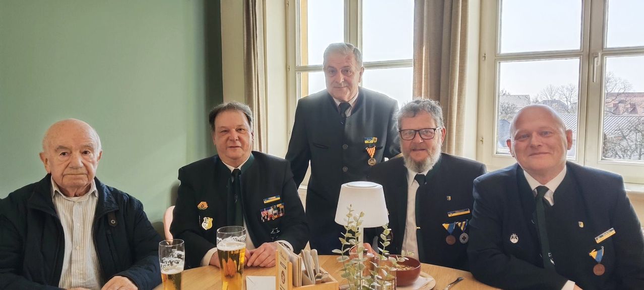 Drei Männer in Uniformen und Krawatten sitzen an einem Tisch mit Getränken und einer Pflanze. Der Mann links lächelt, der Mann in der Mitte steht und der Mann rechts trägt eine Brille.