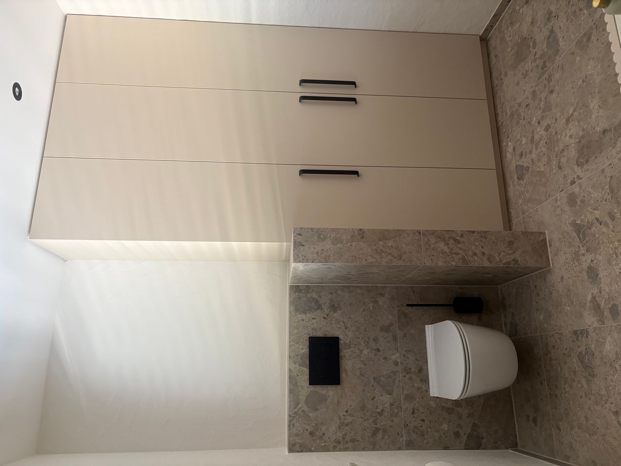 Eine Badezimmerecke mit einem weißen Toilettensitz und einer beigen Wand, ausgestattet mit drei Schubladen mit schwarzen Griffen. Die Wand hat eine Marmorstruktur.