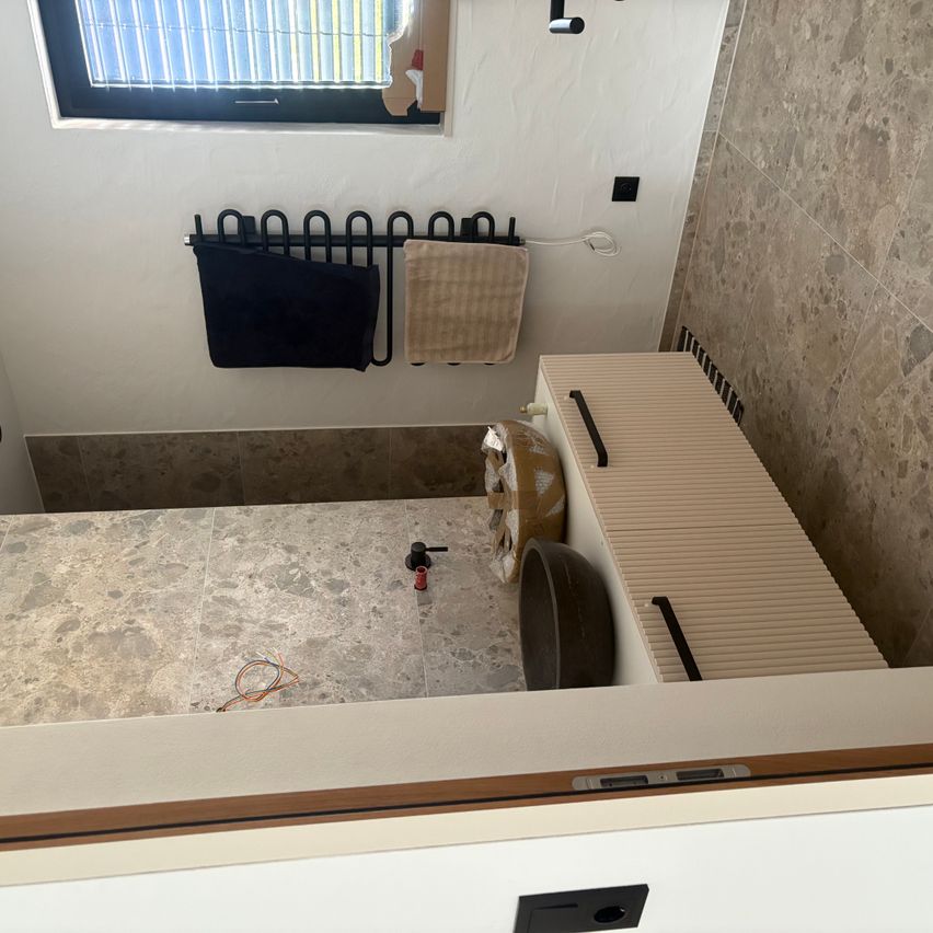 Ein kleines Badezimmer mit Fenster, Handtuchhalter und Waschbecken. Der Boden und die Wände sind gefliest.