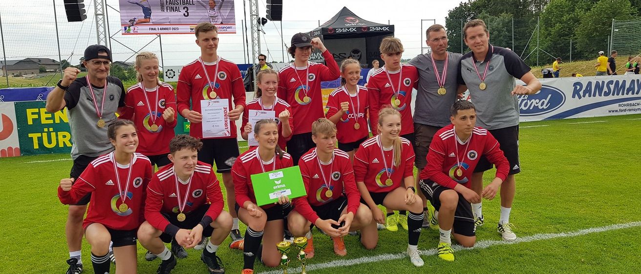 Eine Fußballmannschaft junger Athleten in roten Trikots und Medaillen posiert für ein Foto mit Trainern auf einem grünen Feld. Sie halten Pokale und Urkunden und lächeln in die Kamera.