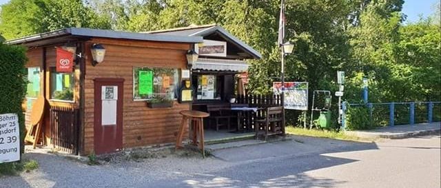 Ein kleines Holzgebäude mit einem Schild an der Tür befindet sich in der Nähe einer Straße, umgeben von Bäumen und Pflanzen. Ein Tisch und Stühle stehen vor dem Gebäude.