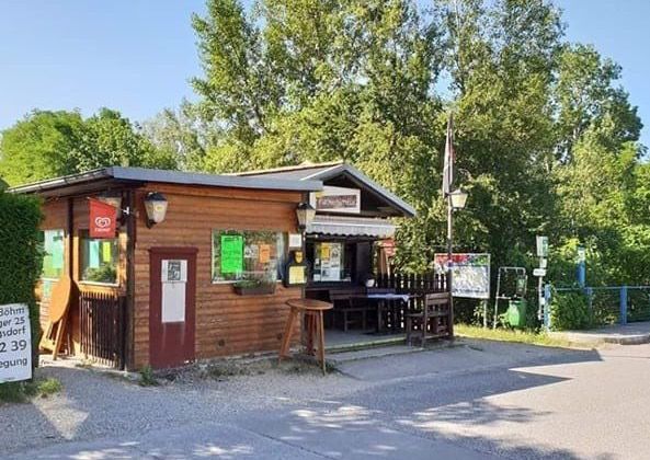 Ein kleines Holzgebäude mit einer Flagge, Tischen und Stühlen draußen, in der Nähe einer Schotterstraße, umgeben von Bäumen und Pflanzen.