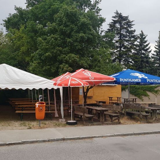 Ein Outdoor-Sitzbereich an der Straße für ein Restaurant mit mehreren Tischen, Bänken und Sonnenschirmen. Ein orangefarbener Mülleimer ist vorhanden. Die Sonnenschirme haben ein rotes und weißes Design und ein blaues Schild. Bäume und ein Gebäude sind in der Ferne.
