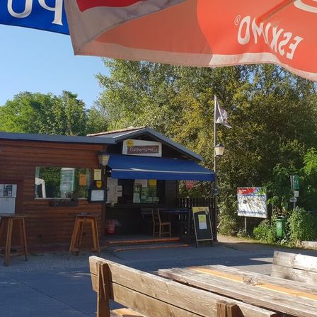 Ein kleiner hölzerner Kiosk mit blauem Vordach, Sitzgelegenheiten im Freien und einem Fahnenmast steht auf einem Kiesplatz mit einem blauen Schild mit der Aufschrift 'Piktobierhütte'. Bäume und Grünflächen umgeben das Gebiet, und eine Holzbank befindet sich im Vordergrund.