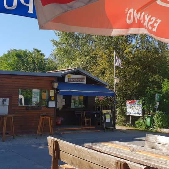 Ein kleiner hölzerner Kiosk mit blauem Vordach, Sitzgelegenheiten im Freien und einem Fahnenmast steht auf einem Kiesplatz mit einem blauen Schild mit der Aufschrift 'Piktobierhütte'. Bäume und Grünflächen umgeben das Gebiet, und eine Holzbank befindet sich im Vordergrund.