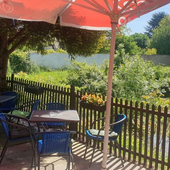 Ein Außenbereich zum Essen mit einem roten Regenschirm, umgeben von einem Holzzaun, Stühlen und Tischen. Es gibt eine Pflanze mit gelben Blumen in einem Pflanzgefäß.