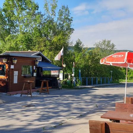 Ein kleines Café befindet sich neben der Straße unter einem roten Regenschirm. Ein Fahnenmast steht daneben und mehrere Bänke sind davor angeordnet. Bäume und ein Zaun befinden sich im Hintergrund.