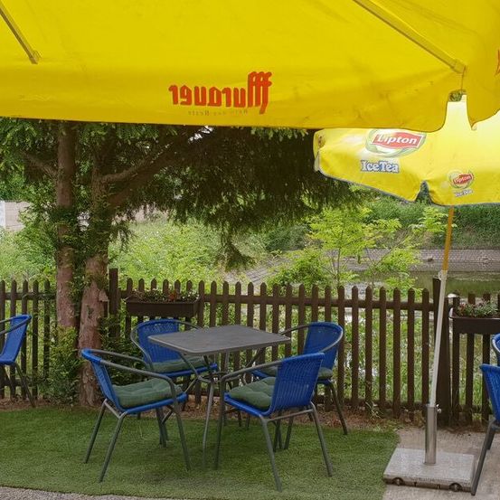 Eine Außensitzecke mit blauen Stühlen und Tischen unter gelben Sonnenschirmen, in der Nähe eines Holzzauns und eines Baumes mit Topfpflanzen.