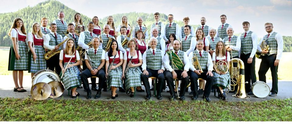 Eine große Gruppe von Musikern, gekleidet in traditioneller deutscher Tracht, posiert mit ihren Instrumenten für ein Foto in einer malerischen Umgebung.