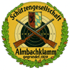 Ein gelbes kreisförmiges Emblem zeigt zwei gekreuzte Gewehre, einen Schild und Zahlen in einem Zielmuster. Der Name 'Almbachklamm' und 'gegründet, 1929' sind in Schwarz geschrieben. Ein grüner Kranz mit Tannenzapfen umgibt das Emblem.
