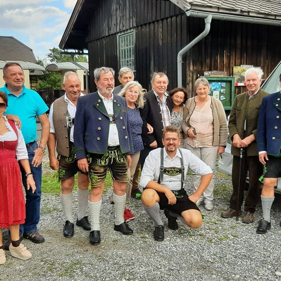 Eine Gruppe von Menschen in traditioneller deutscher Kleidung steht vor einem Gebäude, mit einer Frau in einem roten Kleid und einem Mann in einem blauen Hemd.