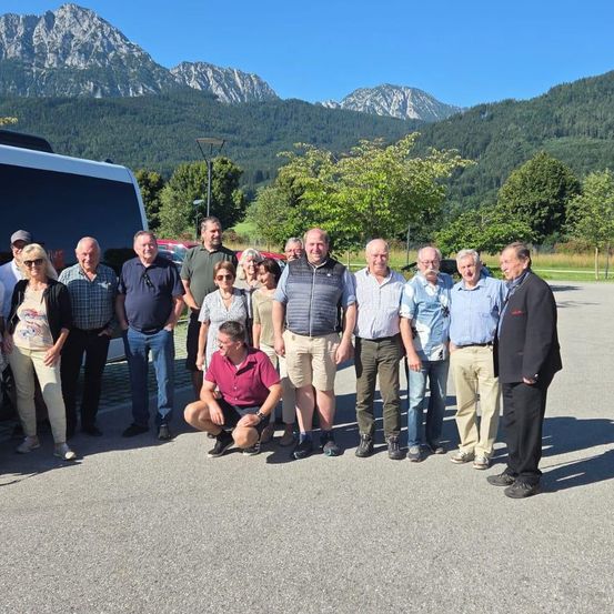 Eine Gruppe von Erwachsenen steht auf einem Parkplatz mit Bergen im Hintergrund. Einige lächeln und posieren für ein Foto.