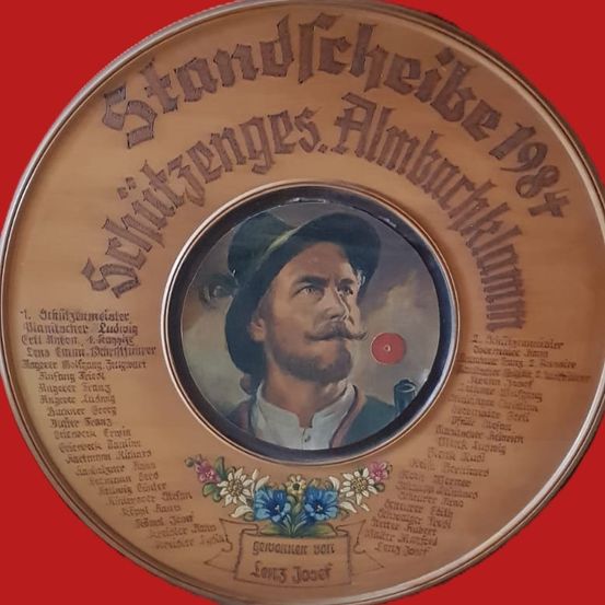 Ein runder Teller mit einem Porträt eines Mannes, der einen Hut und einen Schnurrbart trägt, umgeben von Blumen. Der Teller hat einen roten Hintergrund und beinhaltet Text in Deutsch. Der Text lautet 'Schützenvereins Almbür. 1984'.
