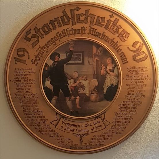Runder Schild mit einem Bild eines Mannes und eines Kindes in einem Raum. Der Schild hat deutschen Text und listet Namen von Personen und Daten auf, möglicherweise ein historischer Bericht oder eine Auszeichnung.