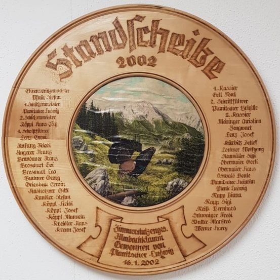 Eine Holzplakette aus dem Jahr 2002 zeigt in der Mitte eine Person, die ein Netz hält, umgeben von Text und Namen. Der Hintergrund zeigt eine Berglandschaft. Der Text und die Namen sind in deutscher Sprache.