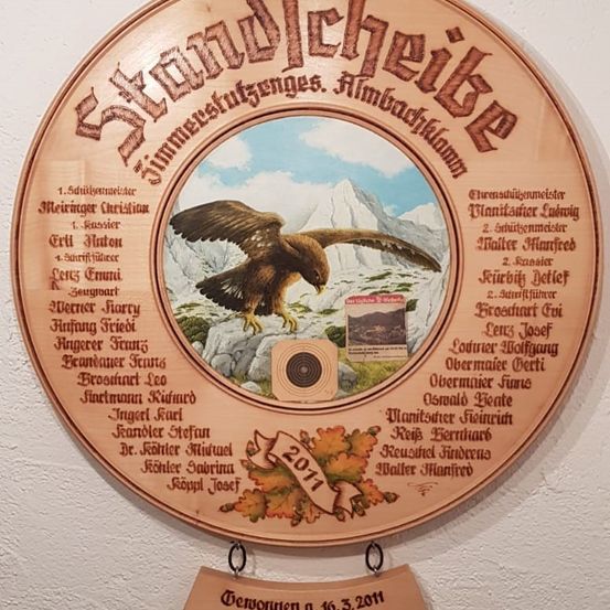 Eine Holztafel mit einem goldenen Adler und einer Zielscheibe in der Mitte. Es hat die Worte 'Standleisbe' und 'Jimmestufengenges.' Es hat eine Liste von Namen und Titeln an den Seiten. Es hat auch ein Datum von 2011.