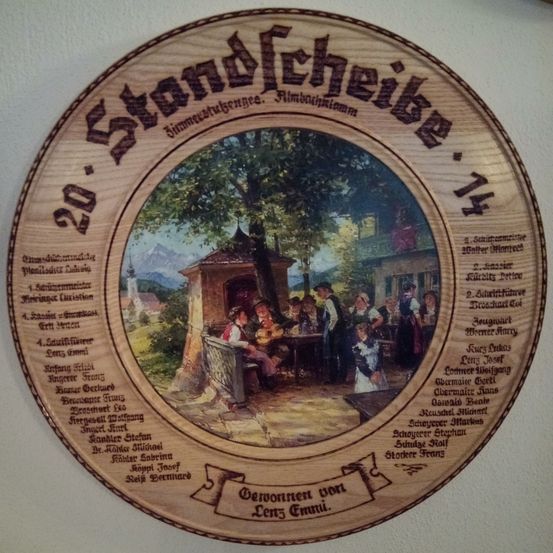 Runde Holzplakette mit einem zentralen Bild von Menschen in einer bayerischen Umgebung. Der Text '20. Standfeche' ist in fetten Buchstaben geschrieben. Die Namen von Personen sind um das zentrale Bild herum eingraviert.