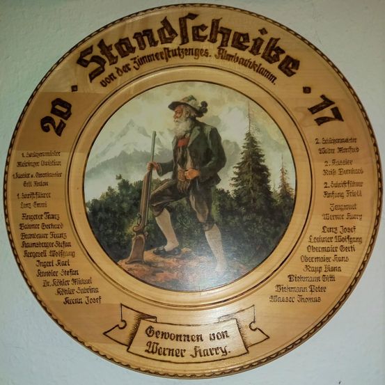 Eine kreisförmige Holzplakette mit einer alten Illustration eines älteren Mannes in traditioneller Kleidung, der ein Gewehr hält, umgeben von Wald und Bergen. Die Plakette enthält Namen und Titel von Personen, mit einer Widmung am unteren Rand.