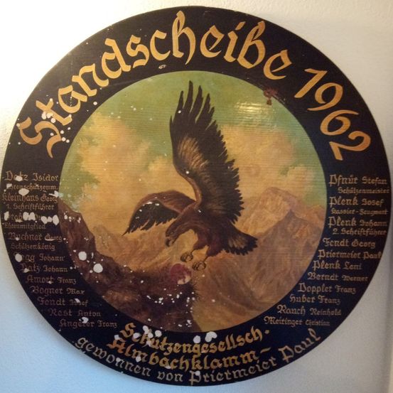 Ein rundes Schild von 1962 mit einer Berglandschaft und einem fliegenden Adler. Namen von Personen sind am Rand eingraviert.