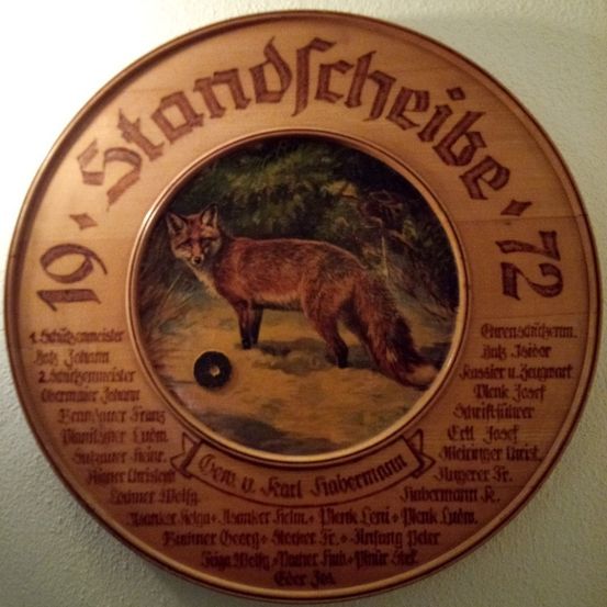 Eine hölzerne Scheibe mit '19 Standscheib'12' und einem Fuchsbild in der Mitte. Namen und Daten umgeben das Bild.