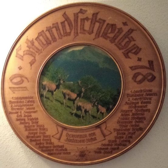 Eine runde Holzplakette zeigt ein Foto von Rehen in einem Feld mit Bergen im Hintergrund. Es trägt den Namen 'Straubing' und verschiedene Texte am Rand.