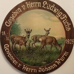 Eine hölzerne Plakette mit kreisförmigem Rahmen zeigt drei Rehe auf einer Wiese. Das Datum 18 XII 1929 ist eingraviert. Die Plakette ist Herrn Ludwig Fuchs und Herrn Johann Wurm gewidmet.