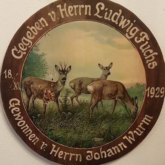 Eine hölzerne Plakette mit kreisförmigem Rahmen zeigt drei Rehe auf einer Wiese. Das Datum 18 XII 1929 ist eingraviert. Die Plakette ist Herrn Ludwig Fuchs und Herrn Johann Wurm gewidmet.