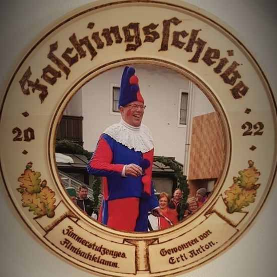 Ein Mann in einem roten und blauen Narrenkostüm ist in einem kreisförmigen Spiegel mit weißem Rand reflektiert. Der Spiegel ist von dekorativem Text umgeben.