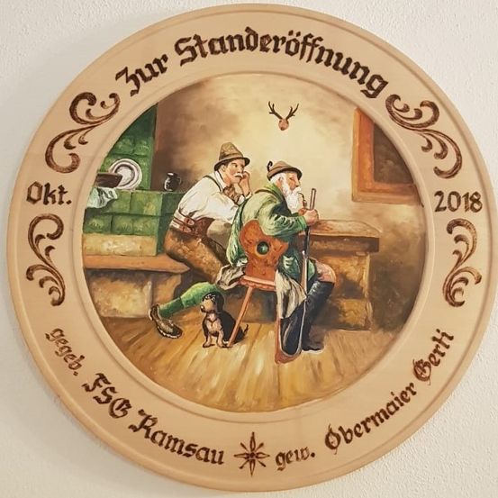 Eine runde Plakette zeigt zwei Männer, die mit einem Hund in einer Taverne sitzen, datiert auf 2018. Der Text lautet 'zur Ständeröffnung'. Ein Mann hält eine Pfeife und einen Stock, der andere ein Bierglas.