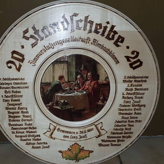 Ein rundes Schild zeigt ein Bild von Menschen um einen Tisch, mit '20. Stadtweihe' oben. Das Bild enthält Namen und Rollen wie 'Erster Meister' und 'Erster Sekretär'. Es hat auch ein Datum '26.2.2020' und Namen von Personen. Das Schild hat einen Rand und ein Blattdesign.