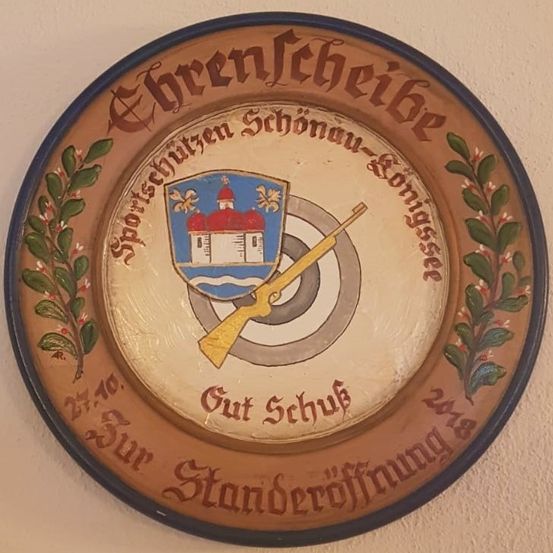 Eine dekorative Holztafel zeigt einen Schild mit einer Kirche und einem Gewehr, umgeben von Blätterwerk. Der Text lautet '27.10. Unser Schützen König 1978 Außer Schutz Standeröffnung.' Sie ist an einer Wand befestigt.