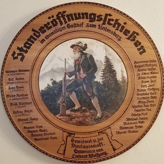 Eine hölzerne Plakette mit deutscher Inschrift, die ein Bild eines älteren Mannes in traditioneller Kleidung mit einem Gewehr vor einem Gebirgshintergrund zeigt. Die Plakette listet Namen und Titel von Personen auf.