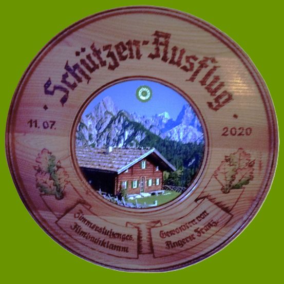 Eine hölzerne kreisförmige Etikette mit dem Text 'Schutzen-Ausflug', datiert auf den 11. Juli 2020. Es zeigt ein Bild einer Hütte mit Bergen im Hintergrund.