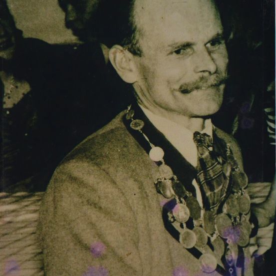 Ein altes Foto zeigt einen älteren Mann mit Schnurrbart, der einen Anzug und eine Krawatte trägt und eine Medaillekette um den Hals hat. Das Bild hat einen Sepiaton.