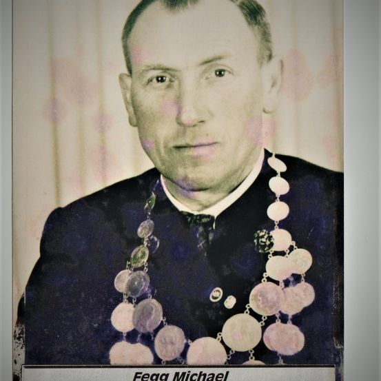 Ein formelles Porträt eines Mannes namens Fegga Michael. Er trägt einen schwarzen Anzug und eine große Halskette mit runden Anhängern. Der Hintergrund ist hell und leicht verschwommen.