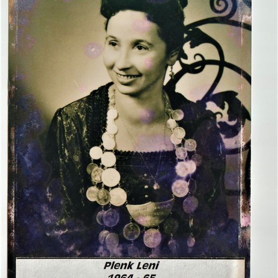 Ein altes Schwarz-Weiß-Foto zeigt eine Frau namens Plenk Leni, lächelnd und mit einer Halskette mit runden Anhängern, aufgenommen 1964.