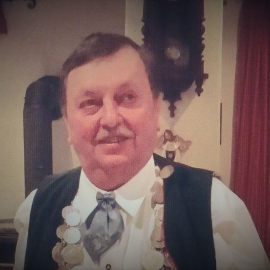 Ein lächelnder Mann in einem weißen Hemd, Fliege und Weste mit Münzen um den Hals steht an einer Wand mit einer Uhr.