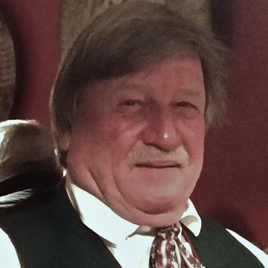 Ein älterer Mann mit grauem Haar und einem Schnurrbart lächelt direkt in die Kamera. Er trägt ein weißes Hemd und eine gemusterte Krawatte.