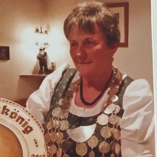 Eine Frau in traditioneller deutscher Kleidung lächelt und steht mit einem runden Teller, auf dem 'Kronung' und das Jahr 2017 steht, vor dem Hintergrund eines Zimmers mit gerahmten Bildern und einem Regal.