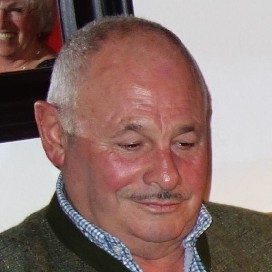 Ein lächelnder Mann mittleren Alters mit Schnurrbart und kahlem Haar trägt ein blau kariertes Hemd und einen braunen Anzug. Er schaut nach links. Hinter ihm lächelt eine Frau in einem roten Oberteil in die Kamera.