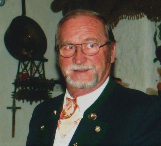 Ein älterer Mann mit Brille und Anzug steht vor einer Wand mit einer hängenden Uhr und einer Pflanze mit roten Blüten.