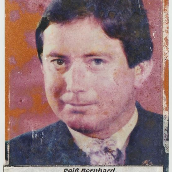 Ein altes Foto eines Mannes namens ReiB Bernhard. Er lächelt und trägt einen Anzug. Der Hintergrund ist leicht verschwommen und rötlich.