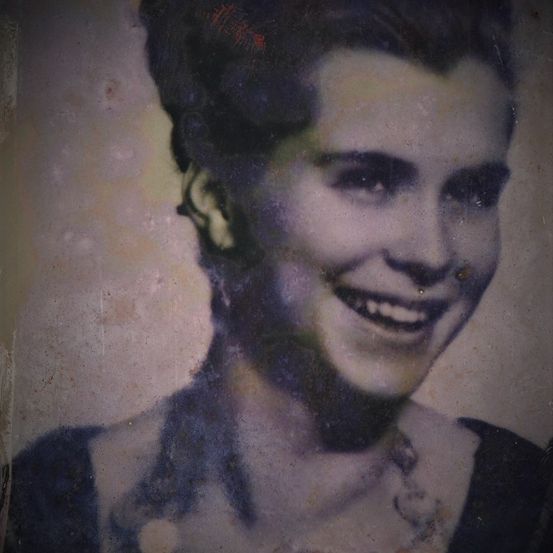 Ein altes Foto zeigt eine Frau mit kurzen Haaren, die ein schwarzes Kleid trägt und lächelt. Das Bild ist leicht verschwommen und hat einen Vintage-Look.