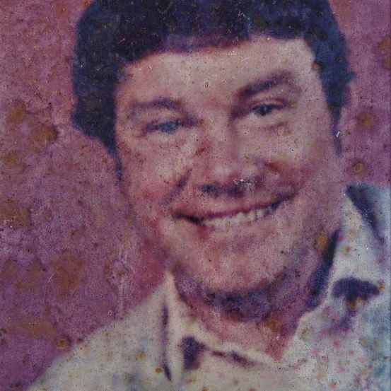 Nahaufnahme eines Mannes mit kurzen dunklen Haaren, der mit einem hellrosa Hintergrund lächelt. Er trägt ein weißes Hemd mit Kragen.