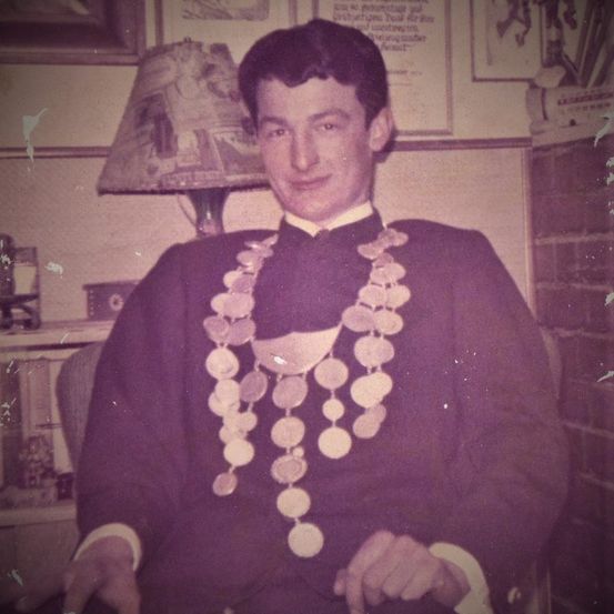 Ein altes Foto eines Mannes, der mit einem lächelnden Gesicht sitzt und eine lange Kette aus Münzen trägt. Hinter ihm ist eine Ziegelwand und ein Regal.