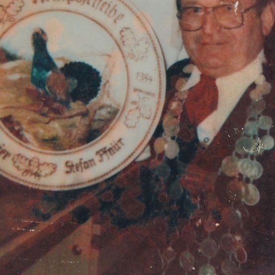 Ein älterer Mann mit Brille hält einen Teller mit einem Vogelbild und dem Text 'Stefan Thun 1984'. Er trägt eine rote Krawatte und eine Girlande.