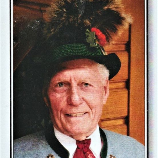 Ein älterer Mann mit einem traditionellen Hut mit einer Feder und einer roten Blume, lächelt für ein Foto. Er trägt einen grauen Anzug und eine rote Krawatte.