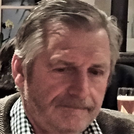 Nahaufnahme eines Mannes mit grauem Haar, der ein kariertes Hemd trägt und ein Glas Bier auf dem Tisch steht.